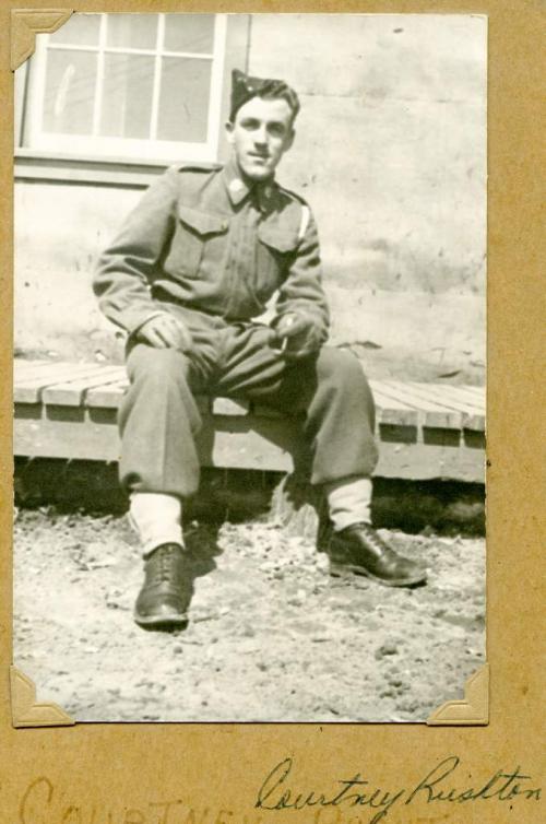 Une photographie en noir et blanc d’un jeune soldat en uniforme assis sur un trottoir de bois.