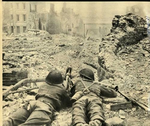 Une photographie en noir et blanc de deux soldats allongés avec un PIAT au bout d’une rue bombardée.