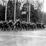 Une photographie en noir et blanc d’un groupe de soldats marchant au pas précédé d’un orchestre militaire de cuivres.