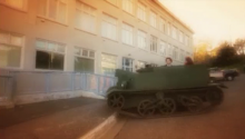 A screen grab of a small army tank with a driver and two people on a street