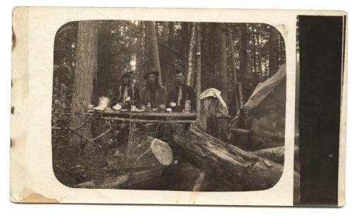 A black-and-white photograph of five men having a meal near a prospector’s tent in the woods.