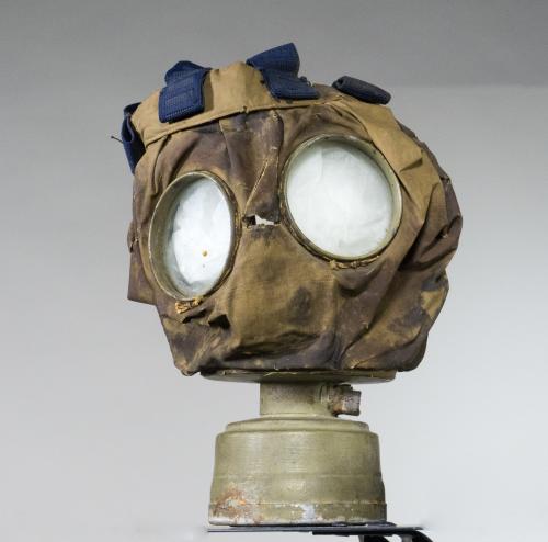 A canvas gas mask with goggle eyes, straps for affixing mask to face, and a  small, metal canister on the bottom.