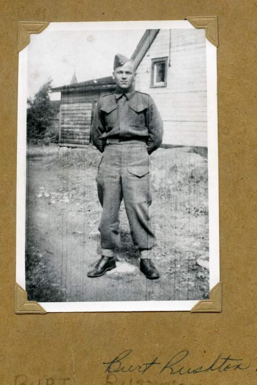 Une photographie en noir et blanc d’un soldat en uniforme posant  devant une maison.