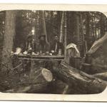 A black-and-white photograph of five men having a meal near a prospector’s tent in the woods.