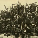 Une image en noir et blanc d’un grand groupe de joyeux soldats canadiens brandissant des obus dans les airs.