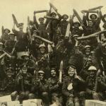 A black-and-white image of a large group of Canadian soldiers happily hoisting   shells in the air.
