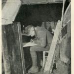 Une photographie en noir et blanc d’un soldat assis dans une tranchée en train d’écrire une lettre.
