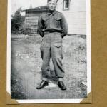 A black-and-white photograph of a soldier in uniform posing in front of a house.