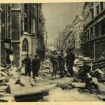 Une photographie en noir et blanc d’un civil et plusieurs soldats dans une ville bombardée.