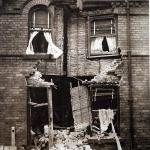 Une photographie d’un bâtiment de deux étages qui a été bombardé et dont la porte d’entrée a été détruite par une bombe.
